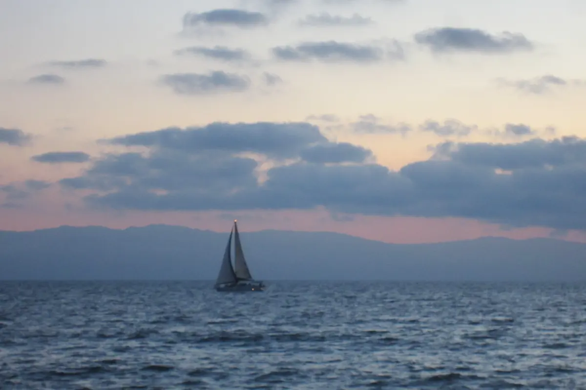 Ein einzelnes Segelboot auf dunkelblauem Wasser unter vereinzelten grauen Wolken, mit dunstiger Bergküste und einem zarten rosa Schimmer in der Dämmerung (ein Segelboot auf der Banderas Bay, kein Showboot)